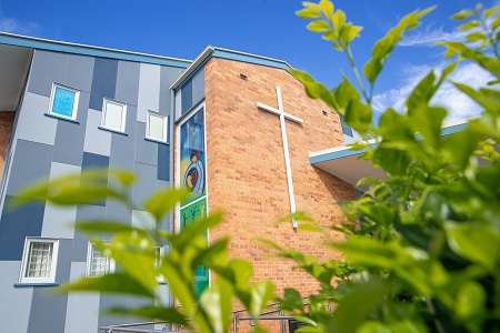 school building with cross feature viewed through greenery highlighting Immaculate Heart Catholic Primary School exterior